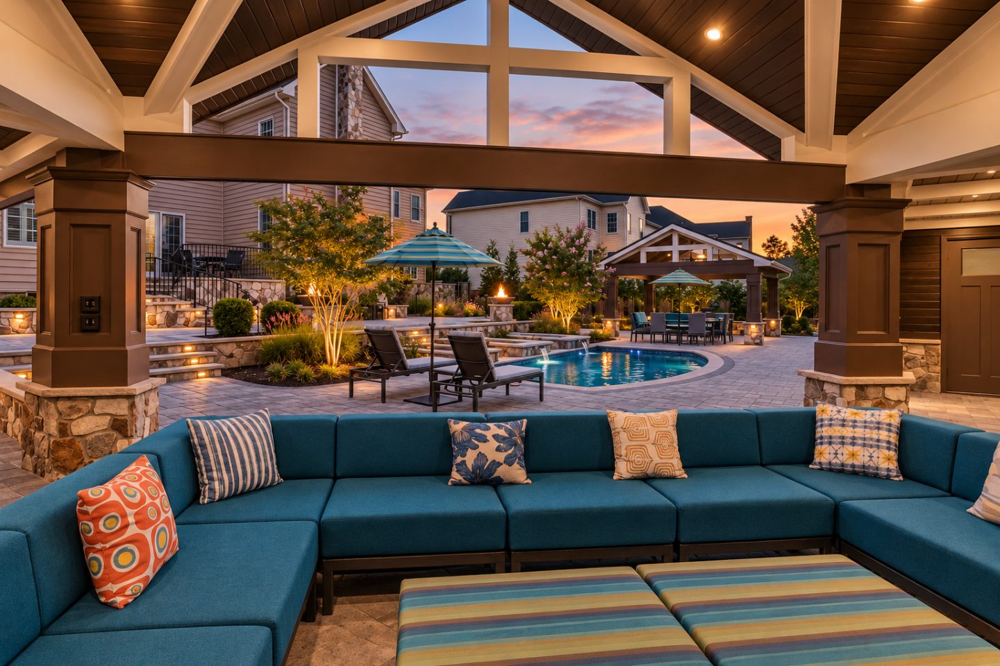 A covered patio with a blue sofa overlooks a lit swimming pool and stone landscaping at dusk.