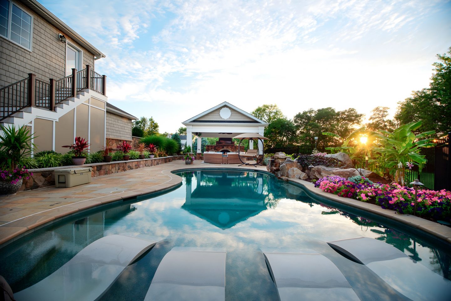 A residential backyard featuring a kidney-shaped pool, stone patio, and landscape lighting fixtures near the plantings at