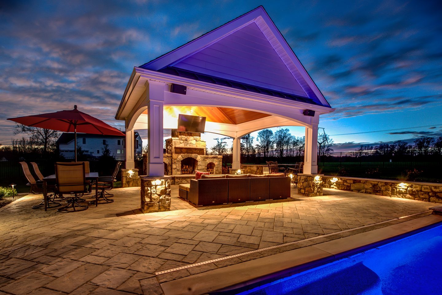 A pavilion with a built-in fireplace sits on a paved patio next to a pool in a residential backyard at dusk.