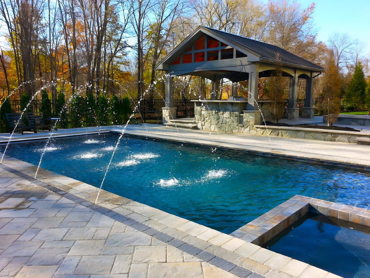 A rectangular pool with fountain jets in a residential backyard featuring a stone patio and covered gazebo
