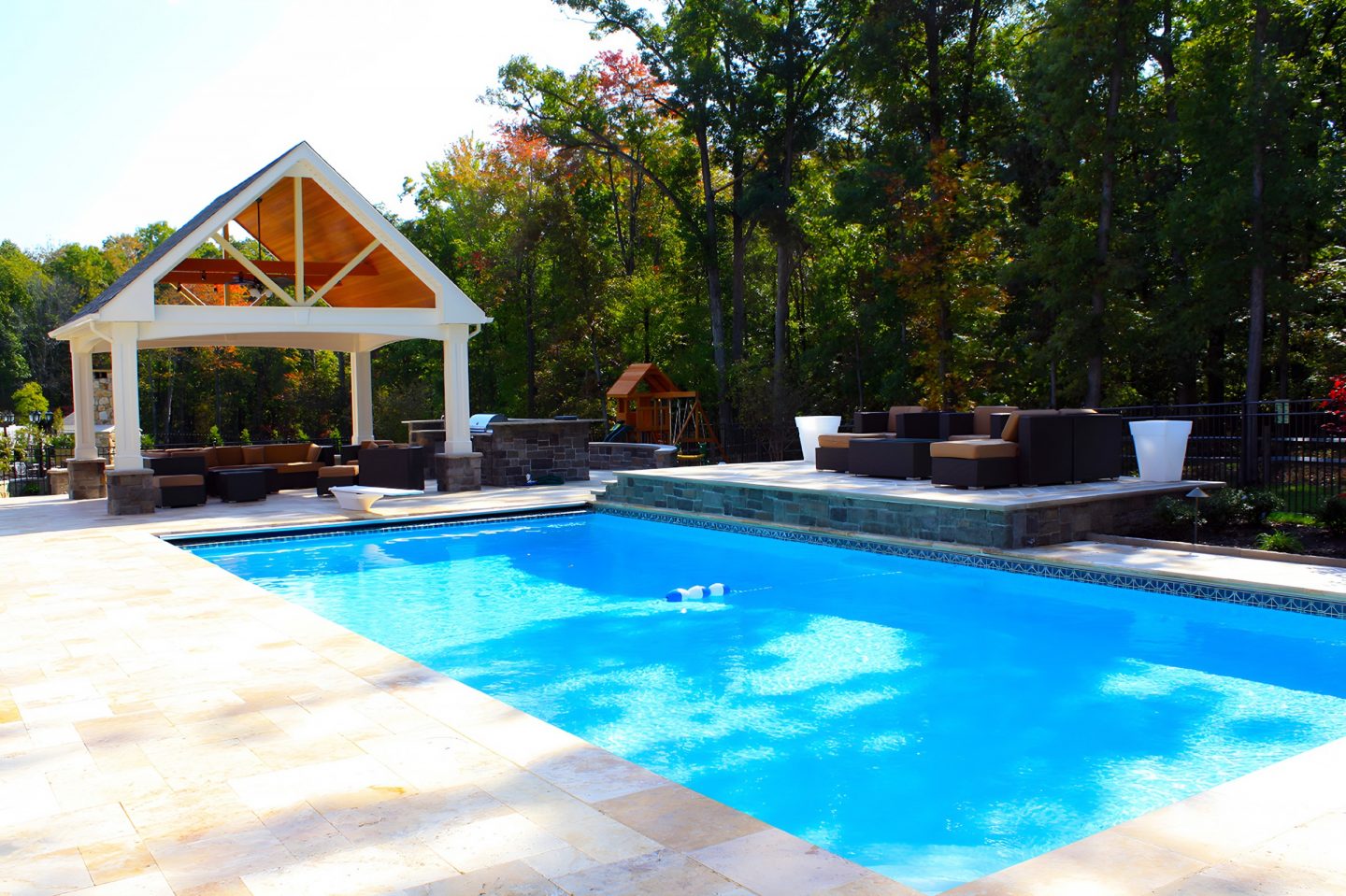 A rectangular swimming pool with blue water is surrounded by a light-colored stone deck and lush green trees.