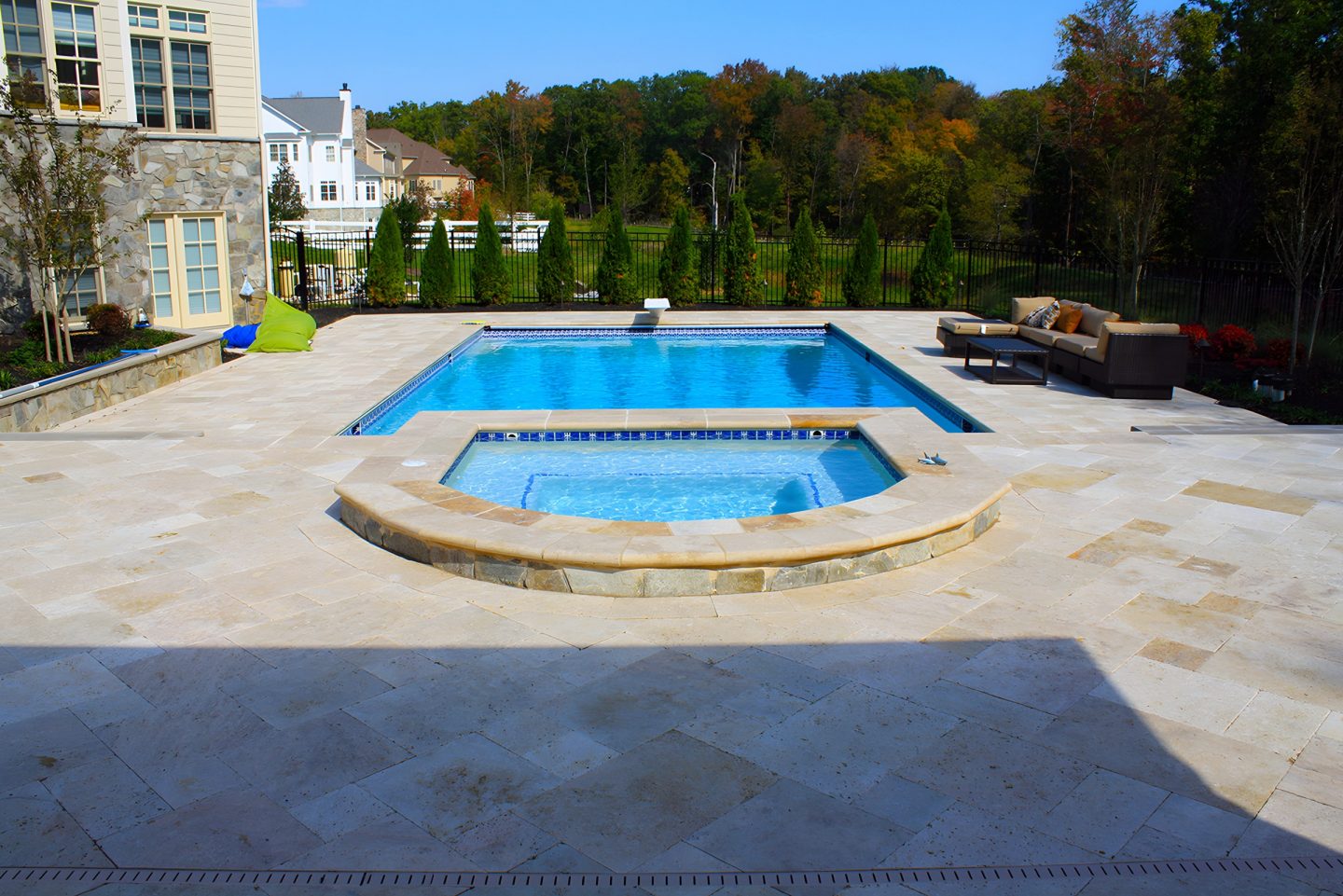 Swimming pool with attached hot tub set into stone patio in residential backyard with autumn trees and outdoor seating