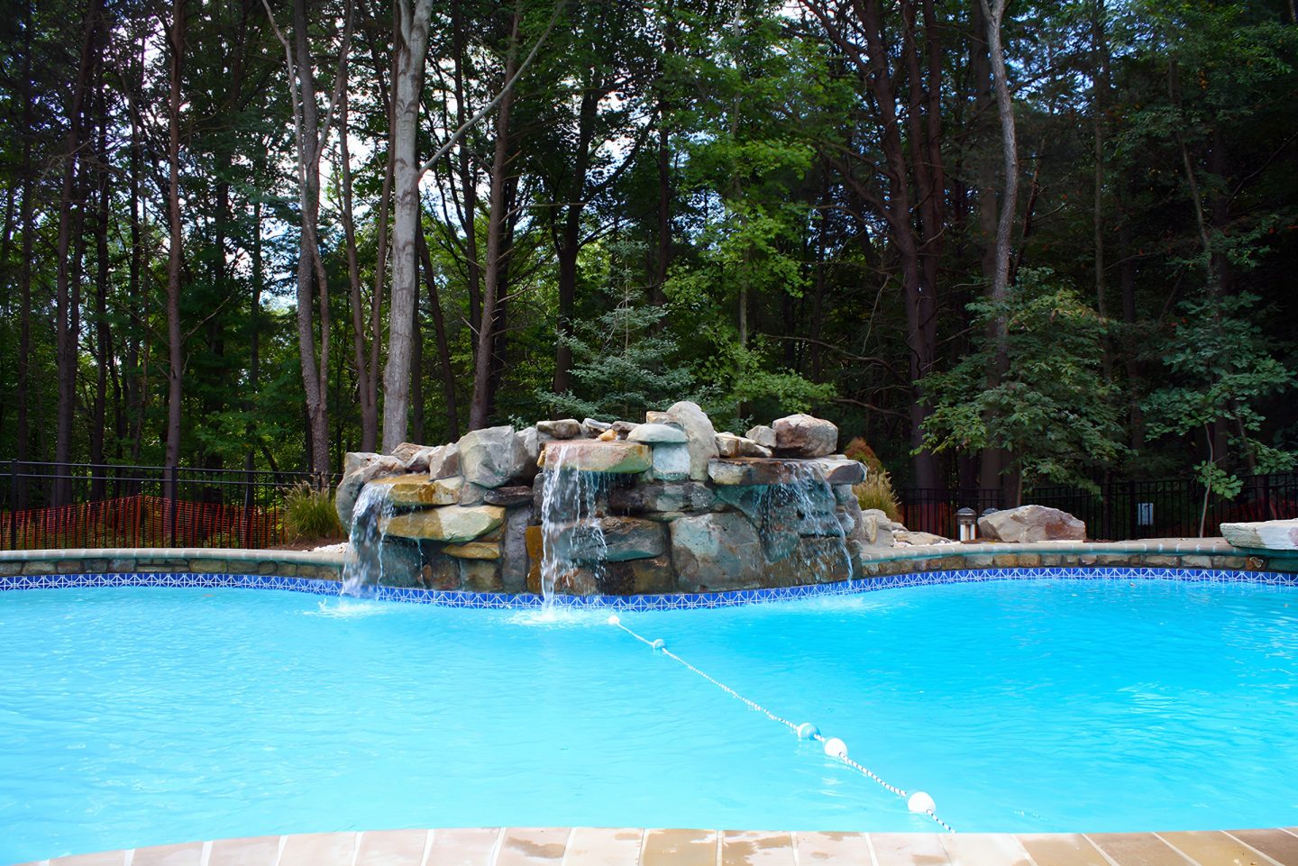 A rock waterfall flows into a pool surrounded by trees and a fence.