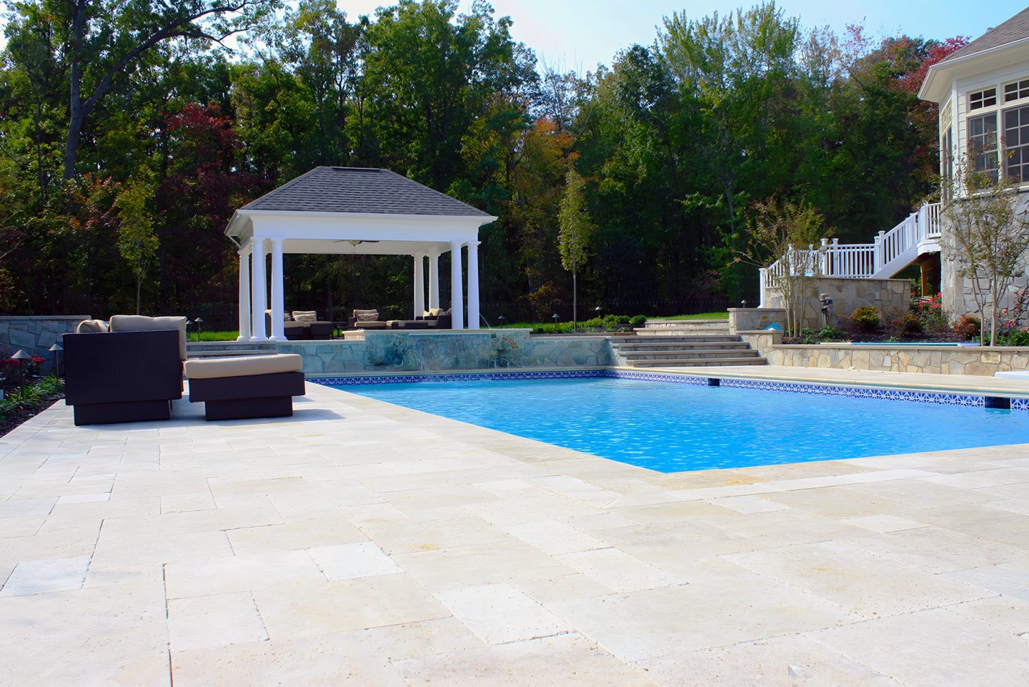 A clear blue pool with a tiled edge in a landscaped backyard featuring a columned gazebo and stone retaining wall.