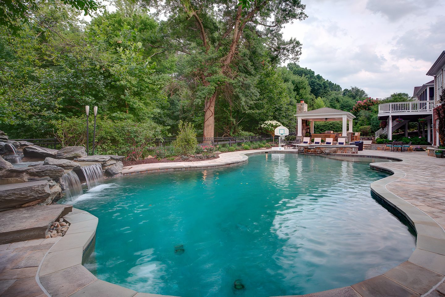 A swimming pool with a rock waterfall in a backyard surrounded by trees and a patio
