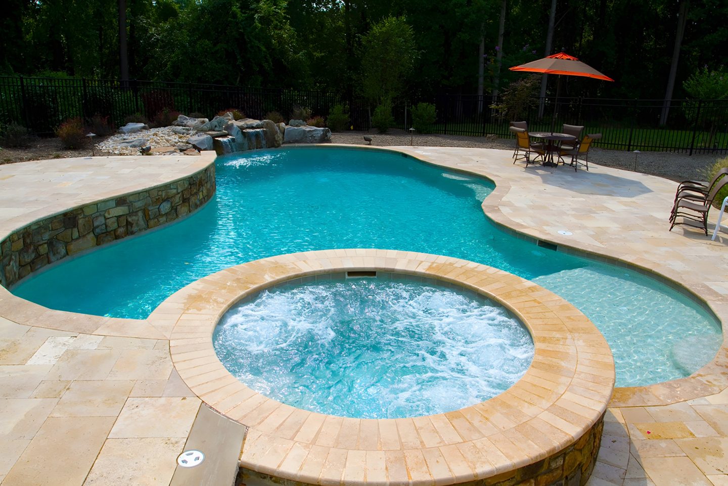 Pool and hot tub in a residential backyard with stone patio and waterfall feature