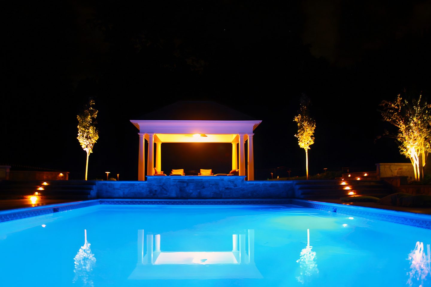Gazebo with columns and cushions sits near a pool at night