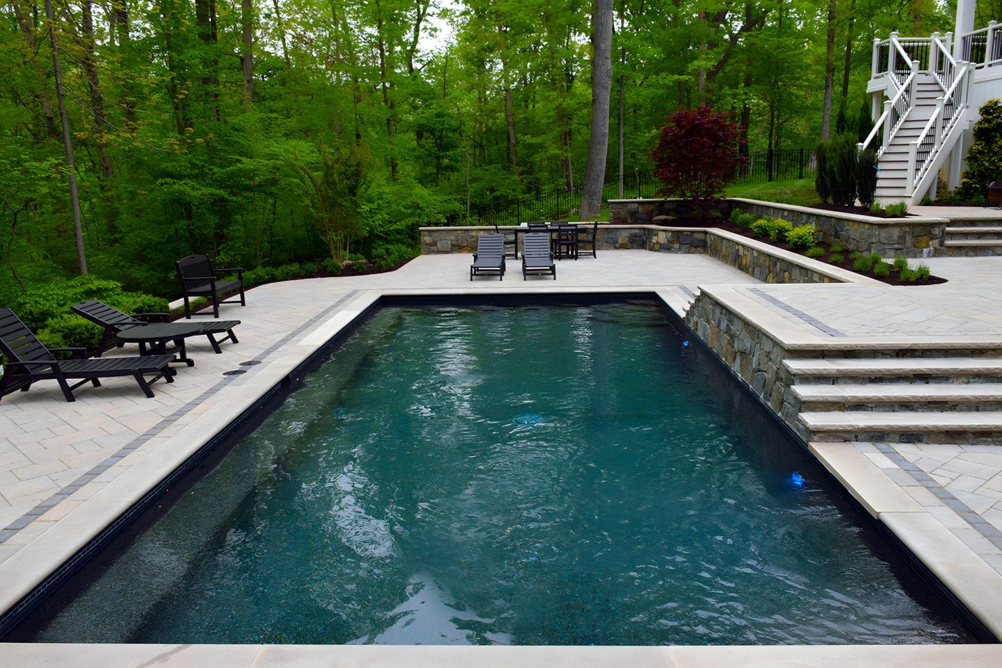 A rectangular pool with blue water sits on a stone patio surrounded by lounge chairs and dining furniture in a backyard