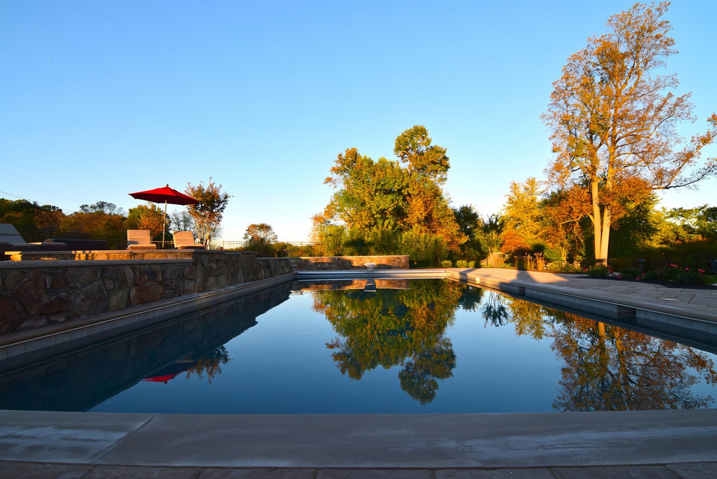A long rectangular pool reflects trees under a clear sky, with a stone wall and seating on the left.