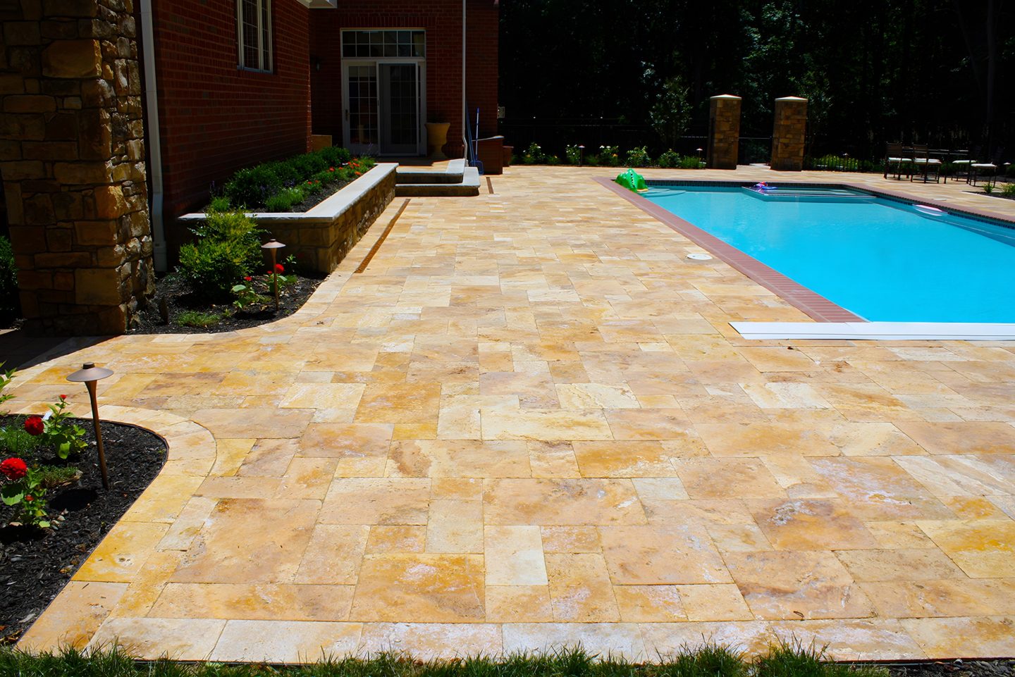 A rectangular pool sits beside a wide patio paved with light brown stone pavers in a staggered pattern.