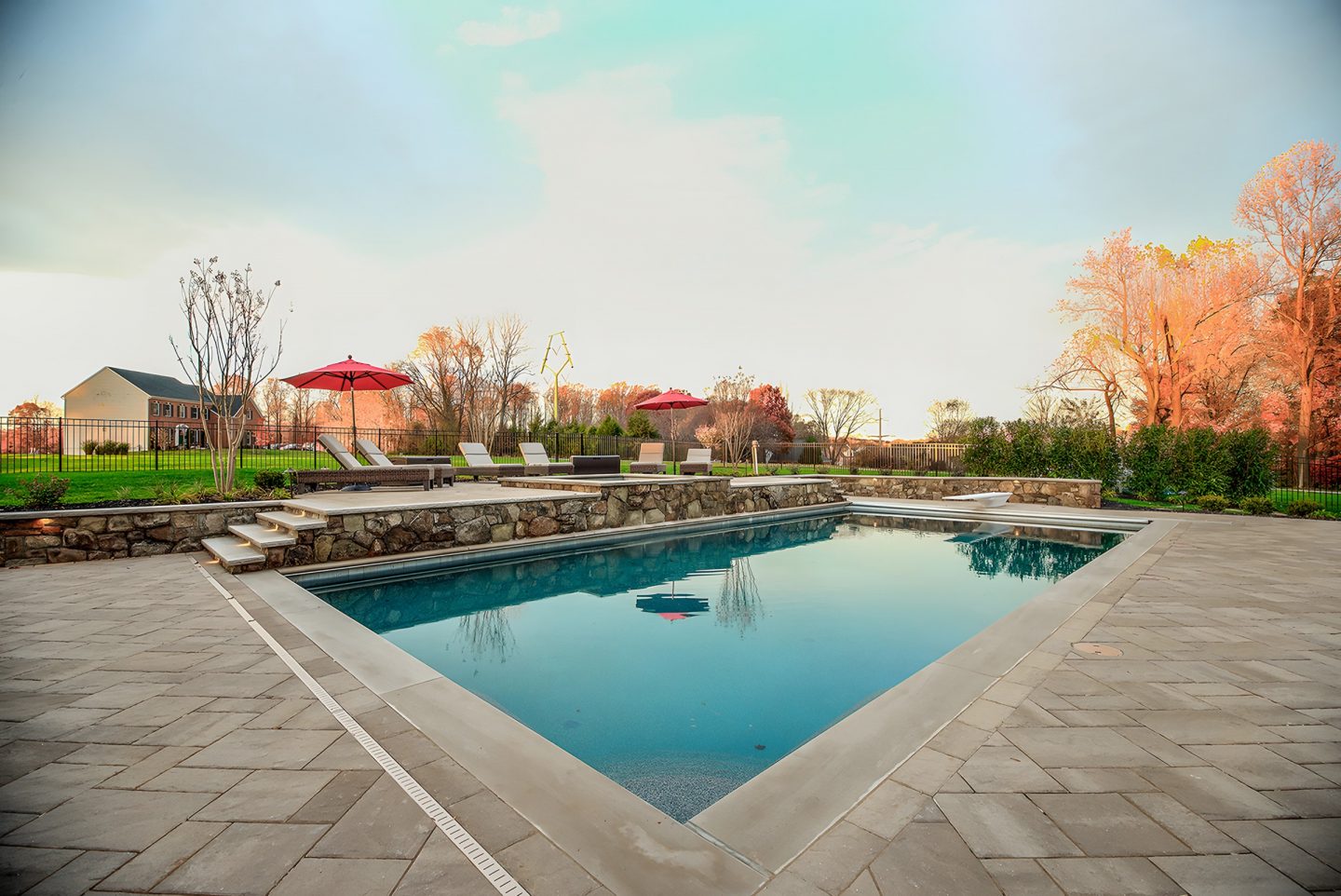 A rectangular swimming pool with clear blue water sits on a stone patio surrounded by steps and red umbrellas in a