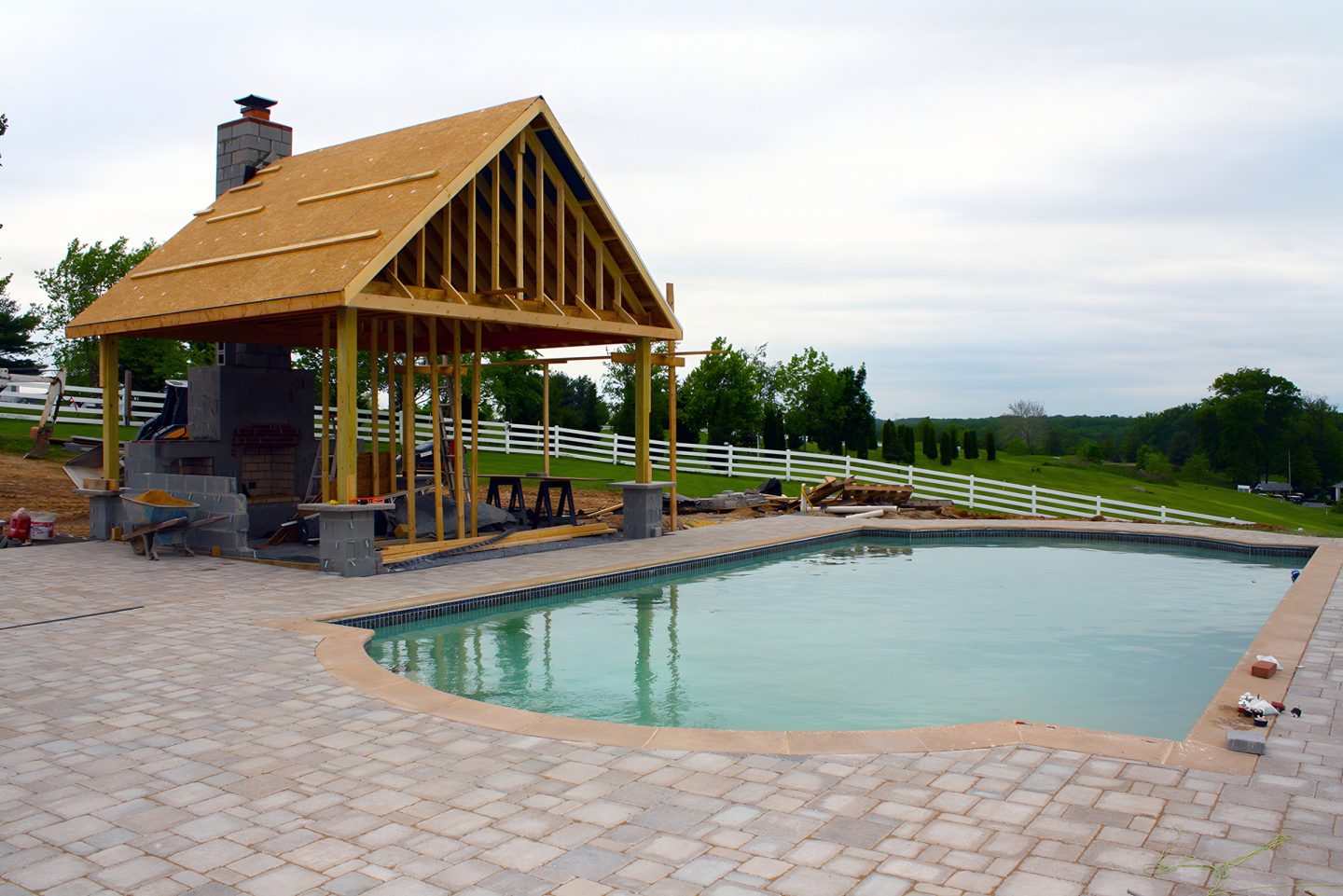 A pergola structure with exposed wood beams stands beside a curved pool edge on a paver deck.