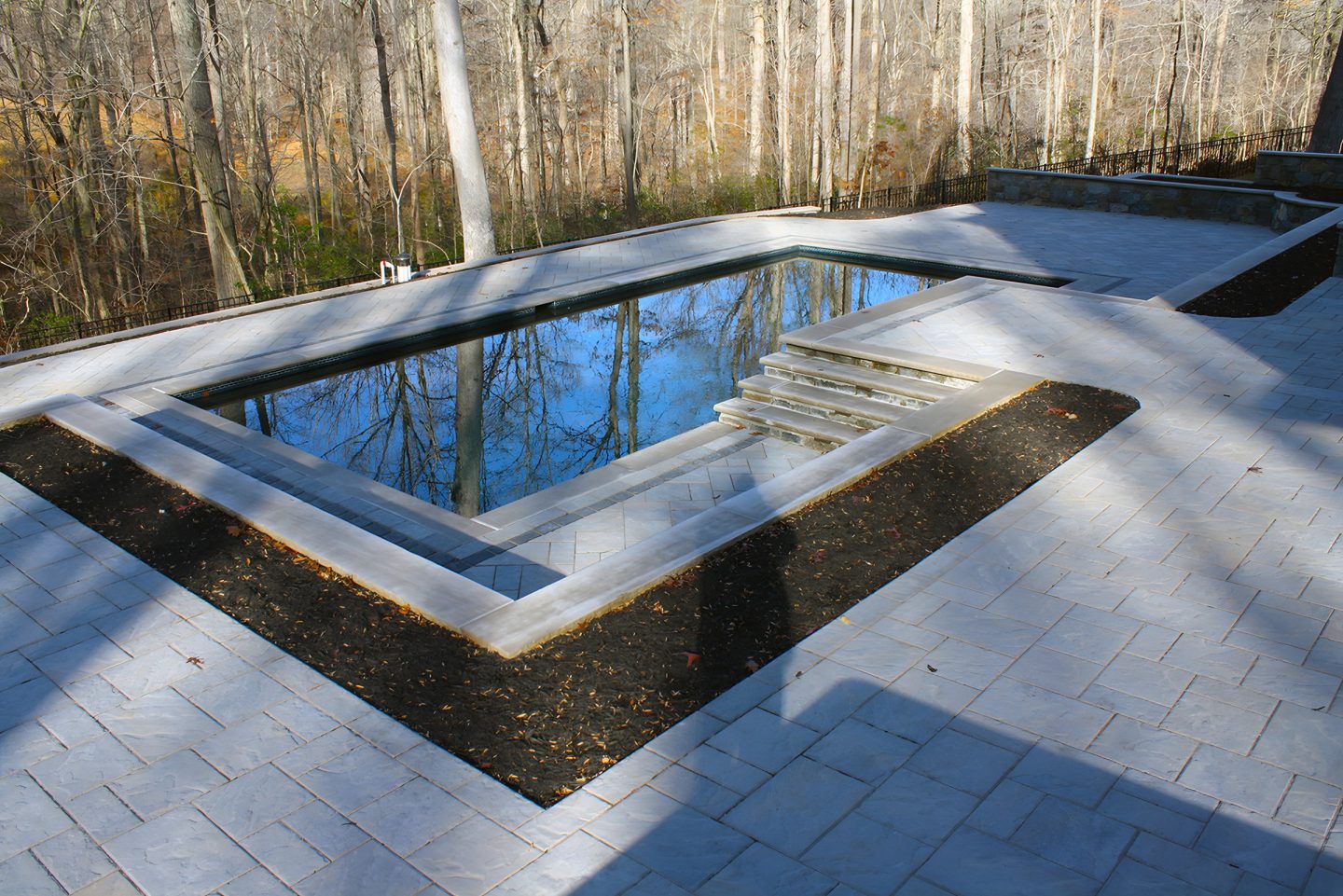 A rectangular pool with steps is set within a paver deck and mulched garden beds in a wooded backyard.