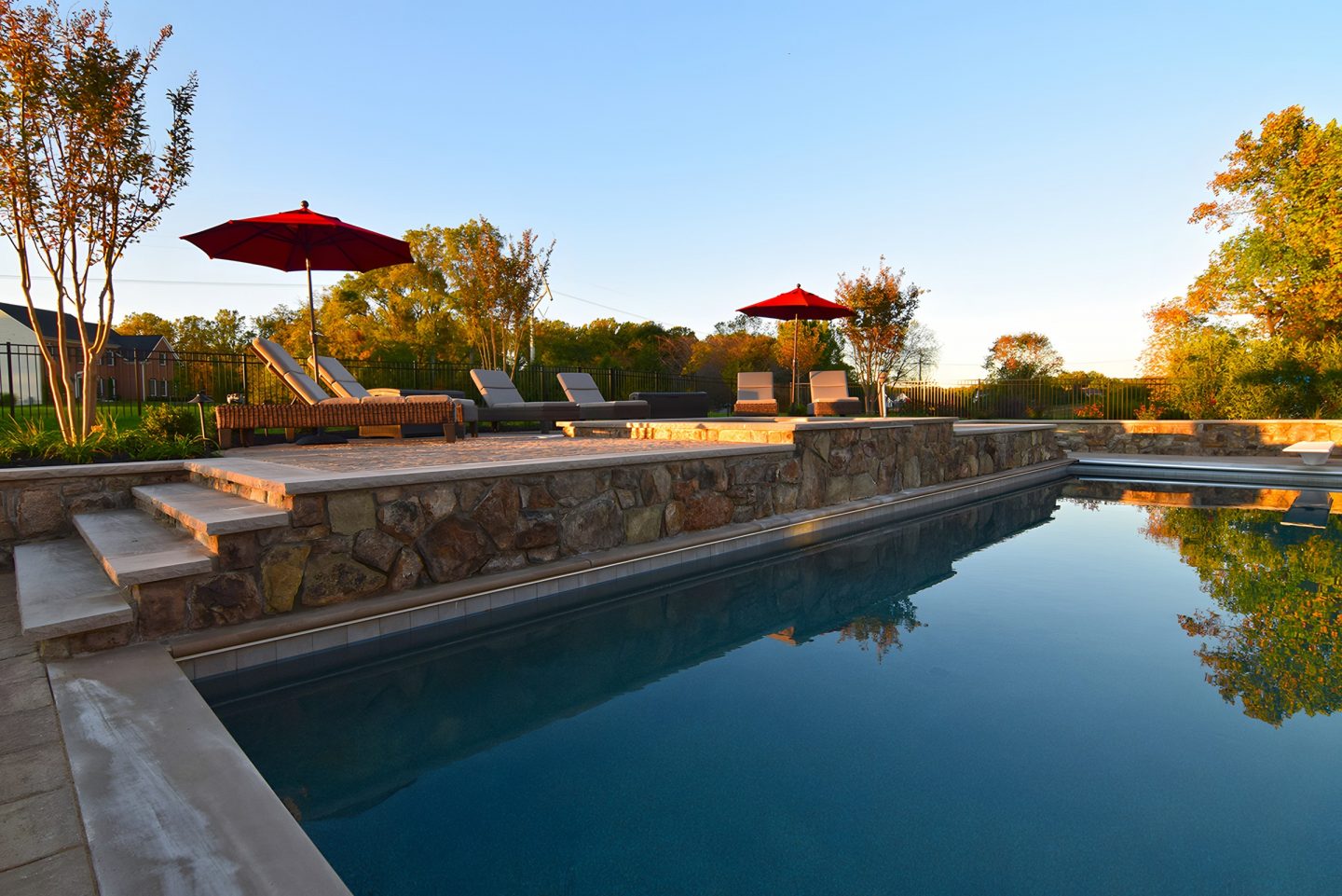 A swimming pool in a backyard with stone walls and lounge chairs under umbrellas