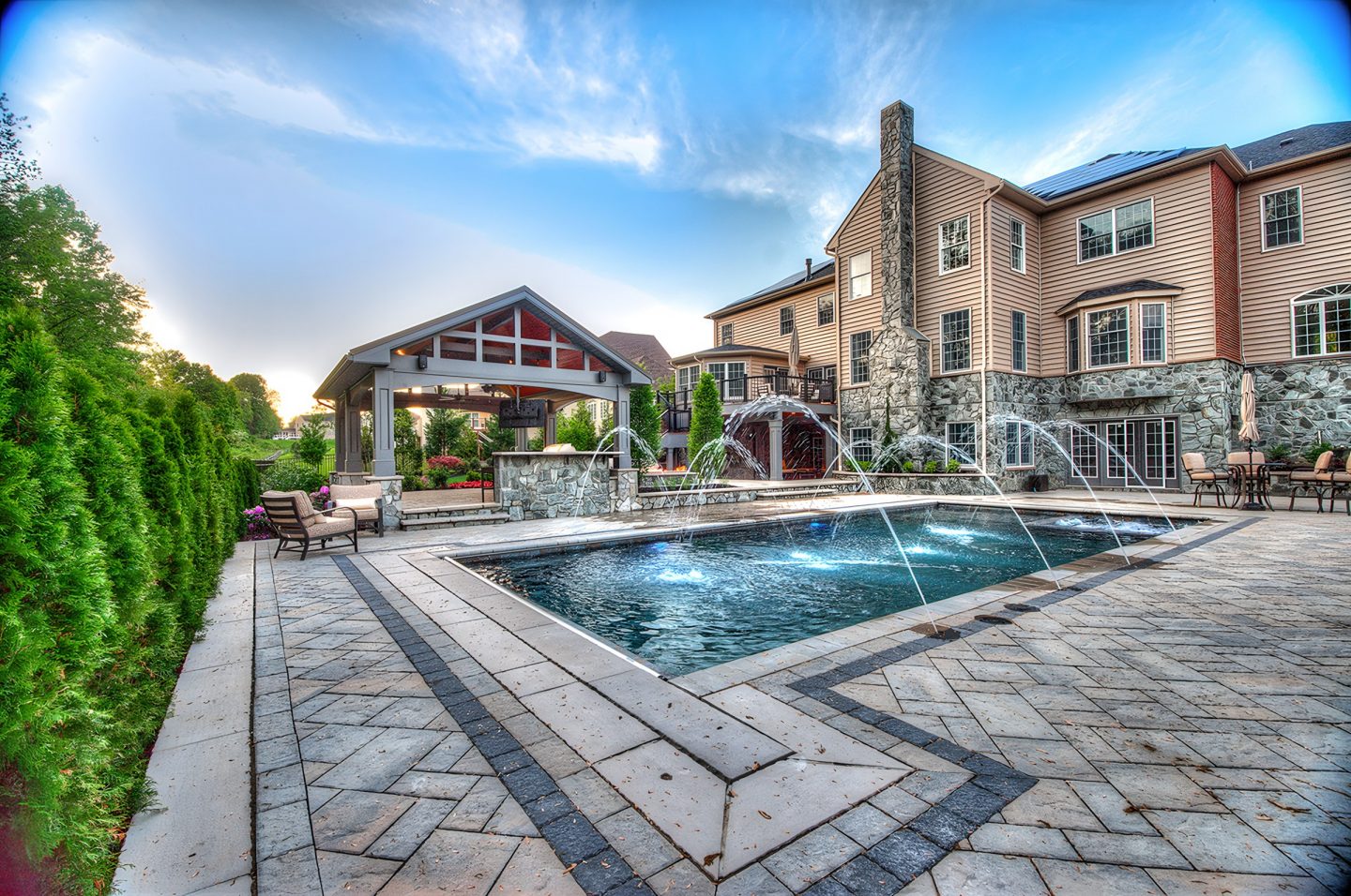 A rectangular pool with water jets is set within a stone patio and landscape architecture.