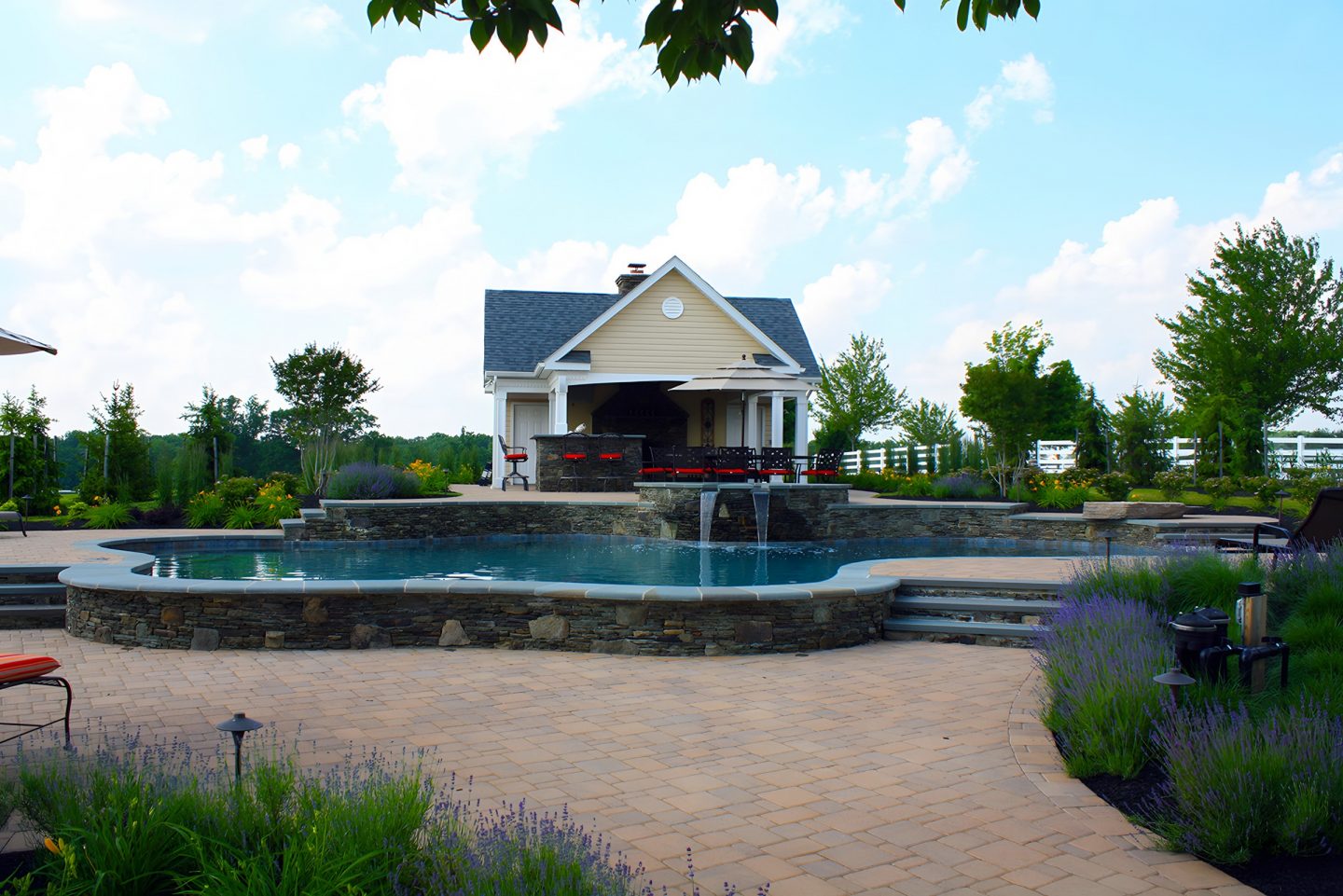 A kidney-shaped swimming pool with a stone coping and waterfall feature sits in a landscaped backyard.