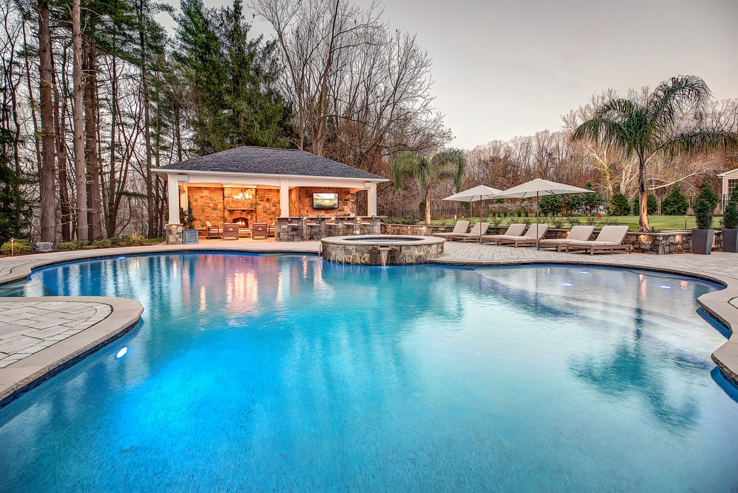 A swimming pool in a residential backyard with a covered pavilion and stone fireplace