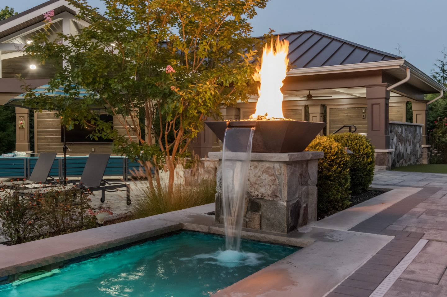 A pool is the central feature in this residential backyard scene at dusk, surrounded by a stone pillar with a flowing