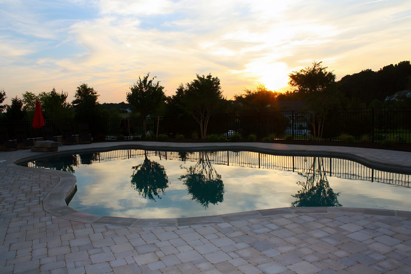 A kidney-shaped pool with stone coping sits in a residential backyard at dusk, surrounded by a paver deck and trees