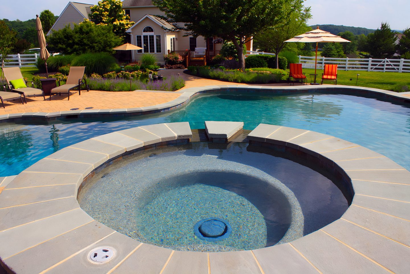 A stone-edged pool with a circular spa feature sits in a landscaped backyard under daylight conditions.