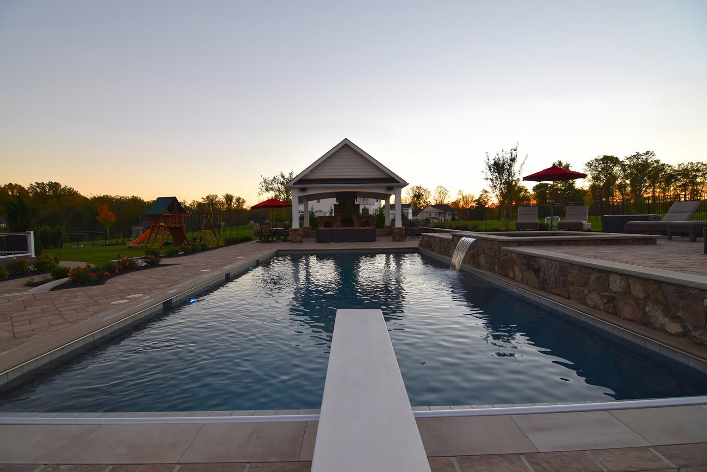 Pool with diving board and waterfall in residential backyard at dusk
